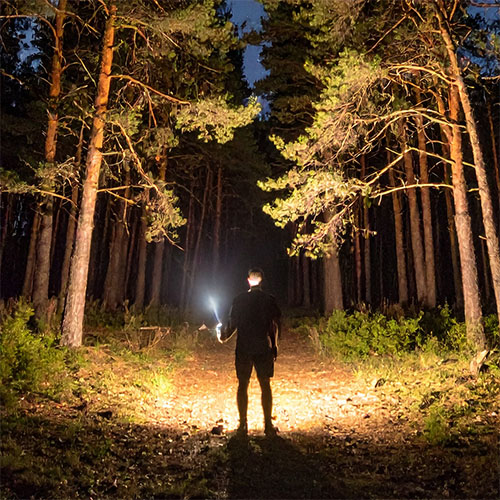 Man standing in a forest at night, lighting the path ahead with a powerful tactical flashlight